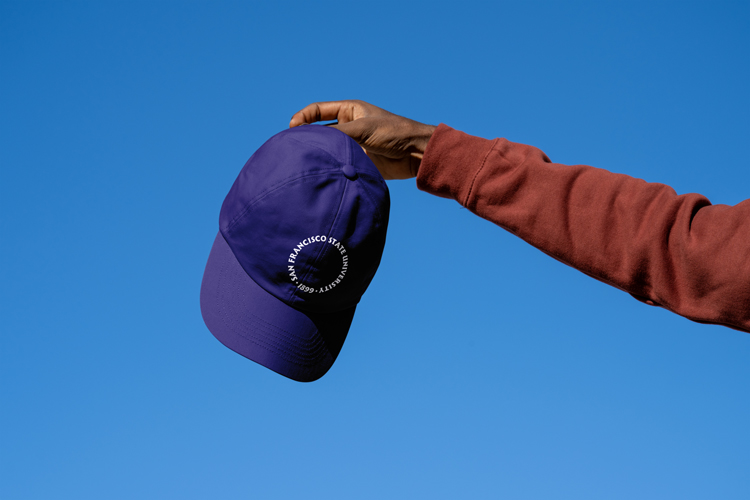 A hand holds up a baseball cap with the SFSU loupe logo printed on the front